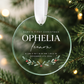 A clear round glass or acrylic baby first Christmas ornament hanging from a white ribbon, printed with the text 'Baby's First Christmas Ophelia Leon' and a small holly leaf design, photographed in front of blurred evergreen branches and warm bokeh lights.