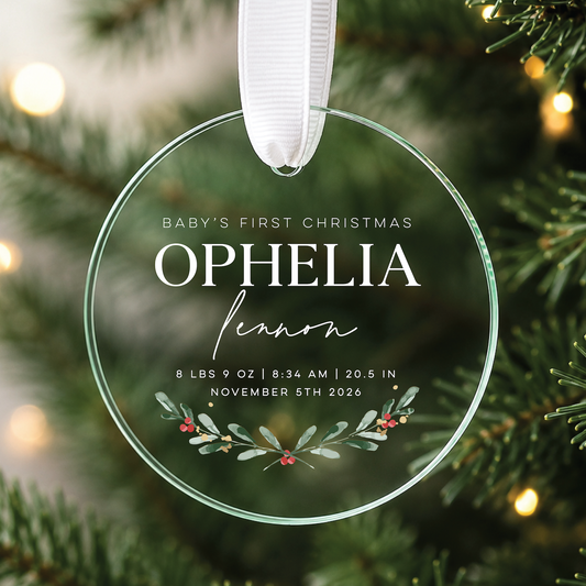 A clear round glass or acrylic baby first Christmas ornament hanging from a white ribbon, printed with the text 'Baby's First Christmas Ophelia Leon' and a small holly leaf design, photographed in front of blurred evergreen branches and warm bokeh lights.