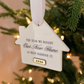 A white house-shaped Christmas ornament hanging from a white ribbon on a Christmas tree. The ornament is printed with the text 'THE YEAR WE BOUGHT Our New Home 65 WEST HARBOUR ST.