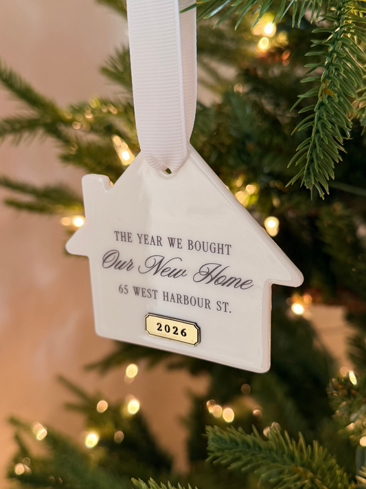 A white house-shaped Christmas ornament hanging from a white ribbon on a Christmas tree. The ornament is printed with the text 'THE YEAR WE BOUGHT Our New Home 65 WEST HARBOUR ST.